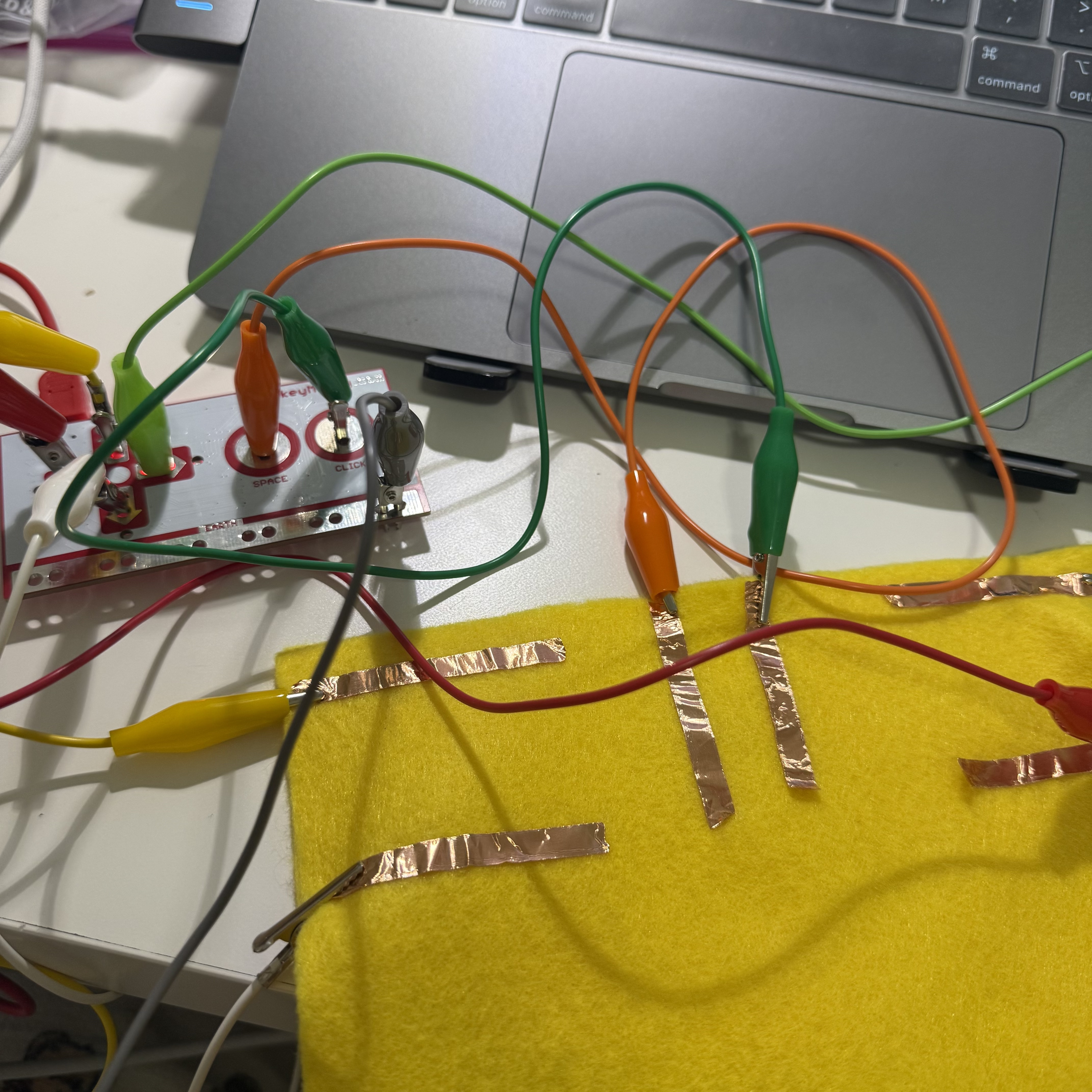 I have set up my Makey Makey board with colorful alligator clips attached to copper strips on a yellow fabric. This configuration is part of an experiment to turn the fabric into a touch-sensitive control surface, allowing me to send commands to my computer through simple touches.